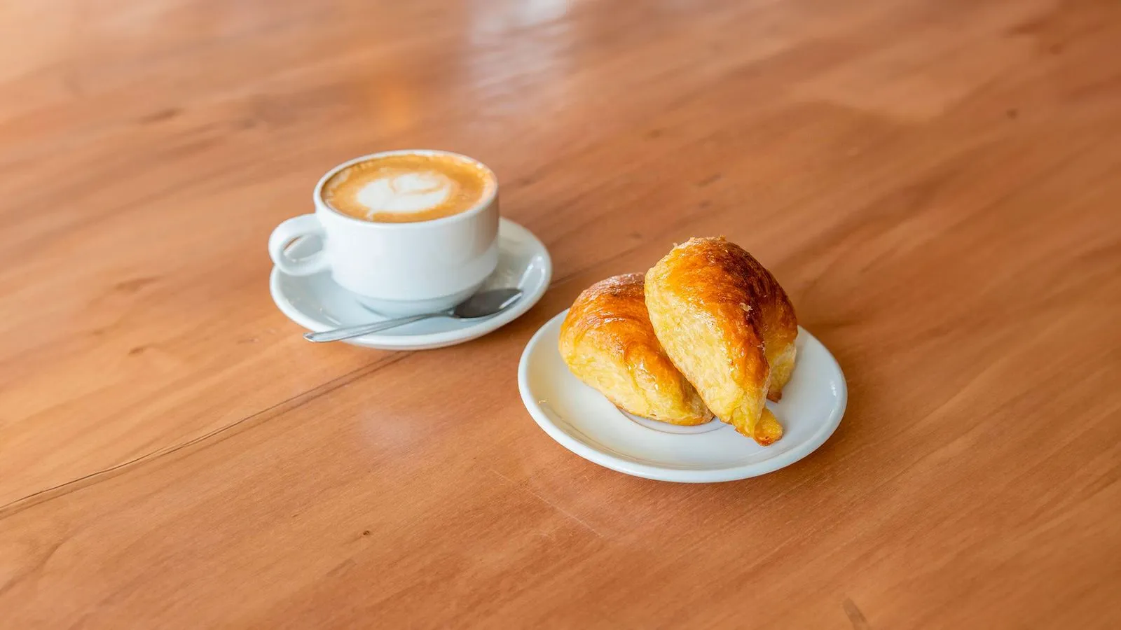 Café con leche con arte en la espuma y dos medialunas sobre una mesa de madera.