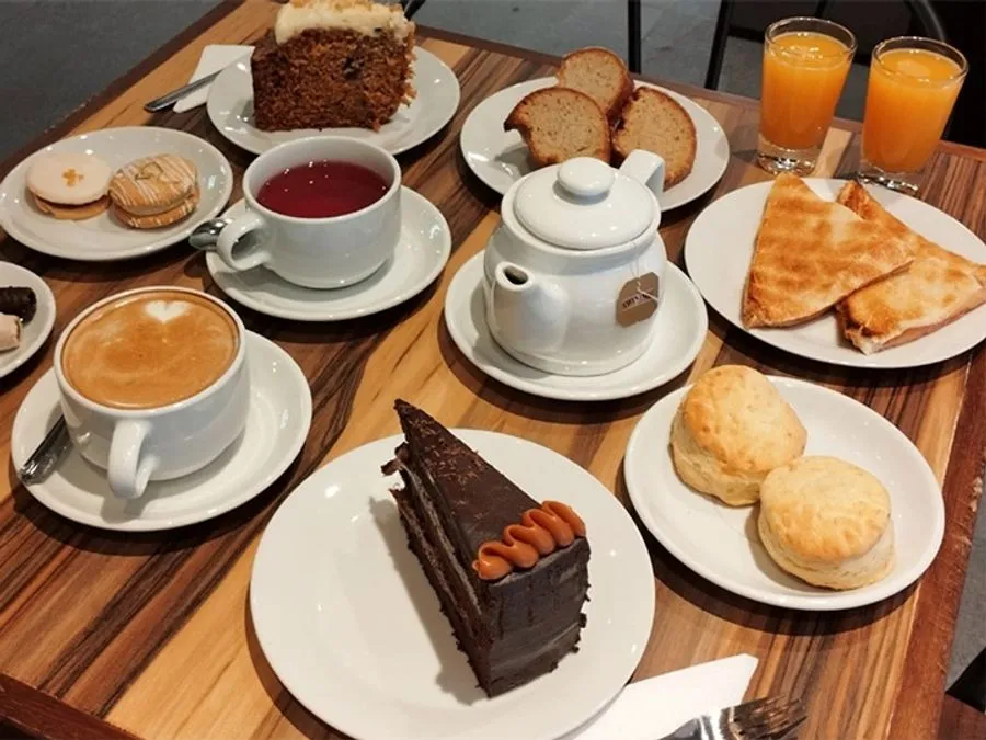 Mesa servida con té para dos: tortas, scones, alfajores, tostados y jugo de naranja.