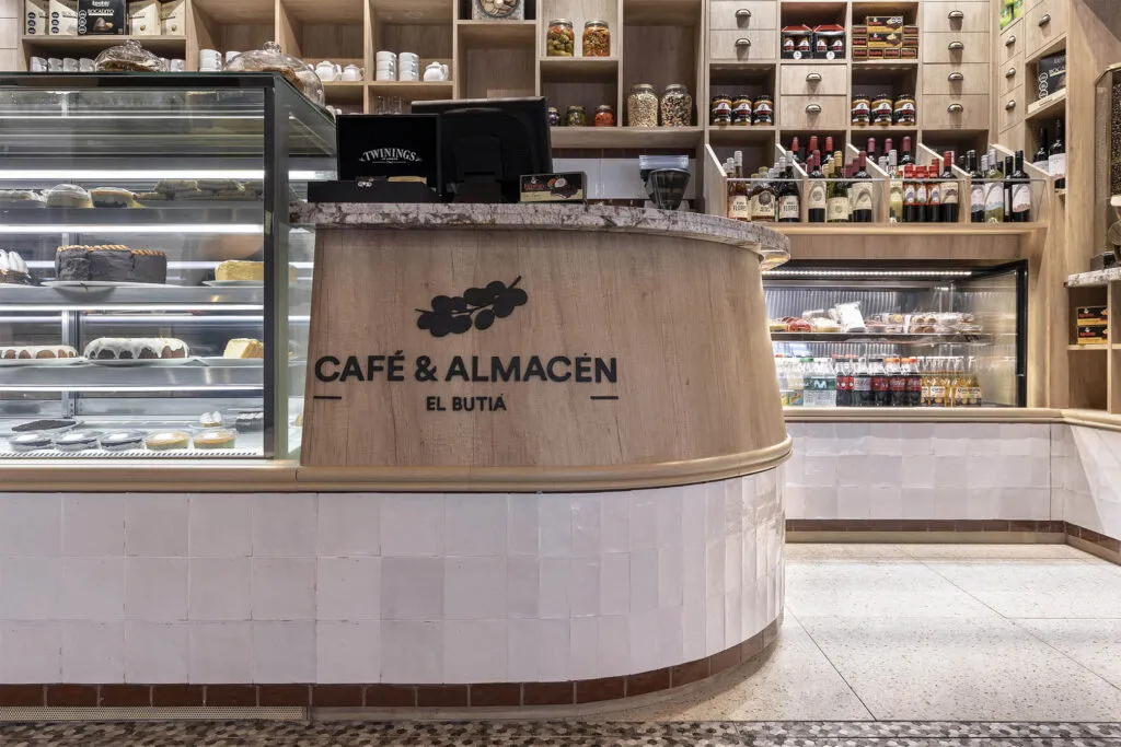 Barra curva de madera del local El Butiá con el logo grabado, vitrina de tortas y estantería de almacén.